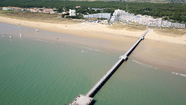 An aerial view of a coastal area with a long pier extending into the sea, a sandy beach, and a cluster of buildings in the background. - Charming 3-BR House w/ WiFi, Near Beach & Forest, Pet-Friendly (Saint-Jean-de-Monts)