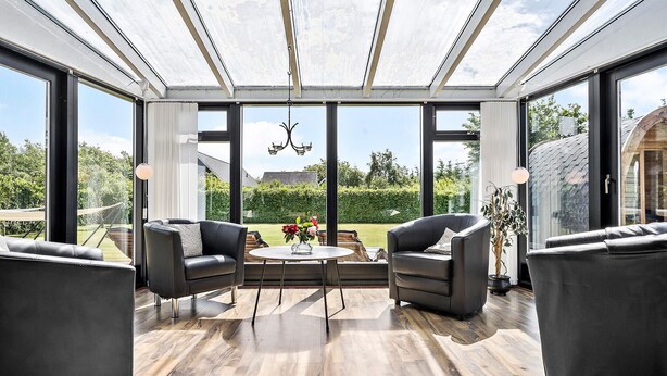 Un salón del vidrio con techo de cristal, dos sofás de cuero, una mesa redonda con flores y vista a un jardín.