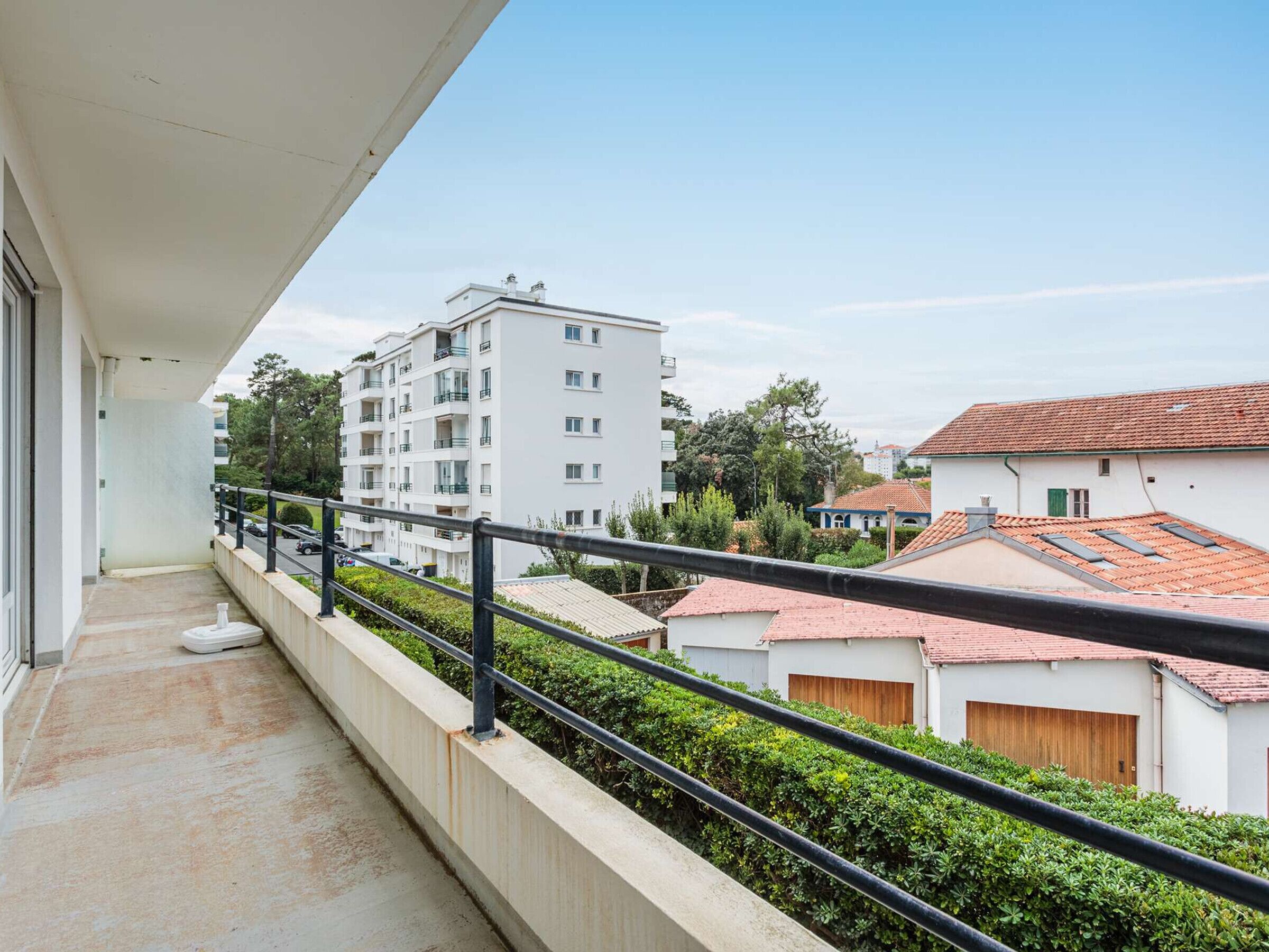 A balcony with a view of a white apartment building, a red-tiled roof house, and a clear sky.