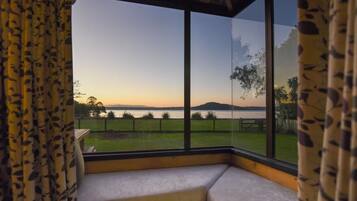 A room with a large window overlooking a grassy area and a body of water during sunset.