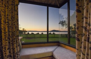 A room with a large window overlooking a grassy area and a body of water during sunset. - Cooper's Lakeside - Lakefront, Kayaks, Sleeps 5 (Rotorua)