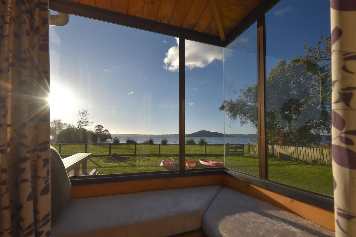 Ein Zimmer mit großem Fenster, das auf eine grüne Fläche mit einer Holzterrasse und Meerblick blickt.