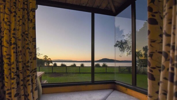 Una habitación con un amplio ventanal que da a un espacio cubierto de hierba y un cuerpo de agua al atardecer.