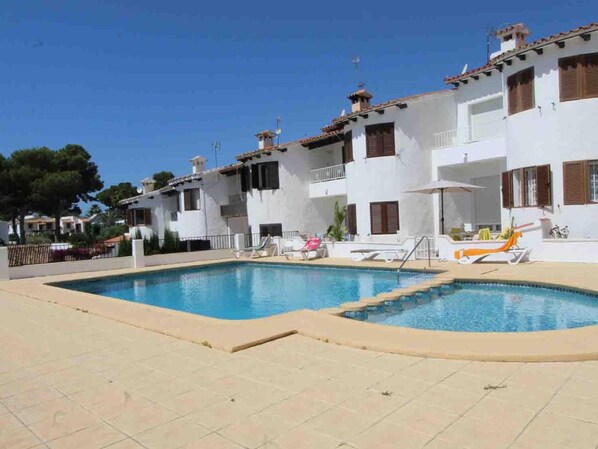 A pool area with a clear blue pool, lounge chairs, and a white building with balconies.