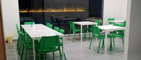A modern dining area with white tables and green chairs, a large window with a view of a cityscape, and a ceiling with recessed lighting.