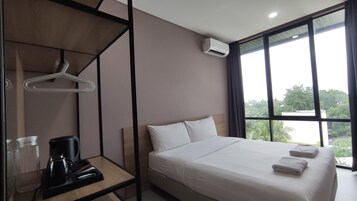 A hotel room with a bed, a desk with a coffee maker, a hanging clothes rack, and a large window with a view of greenery.