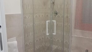 Zona de ducha con mampara de vidrio, pared con azulejos estampados, toallero blanco y panel de control de ducha visible.