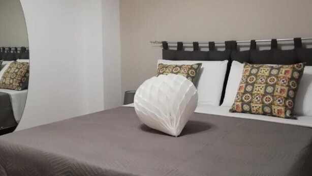 Una cama con almohada blanca con textura y almohadones estampados, cabecera oscura y un espejo en la pared.