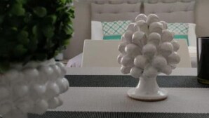 A white decorative vase with spherical elements on a table in a room with a bed and a white headboard.