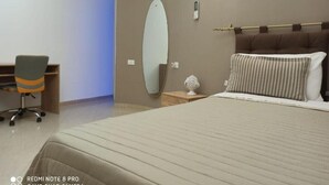 A hotel room with a bed, a desk, a chair, a bedside table with a lamp, and a wall-mounted mirror.