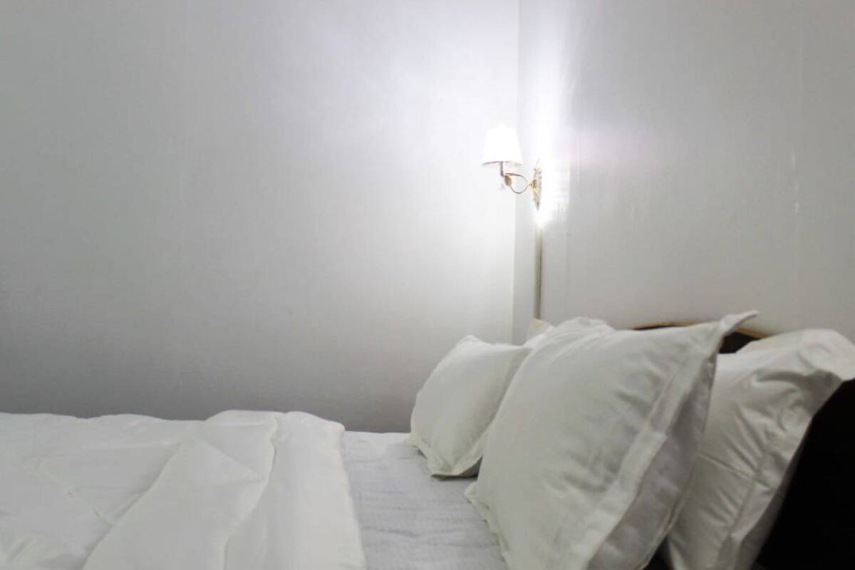 A simple, unadorned bedroom with a bed, white linens, and a wall-mounted light fixture.