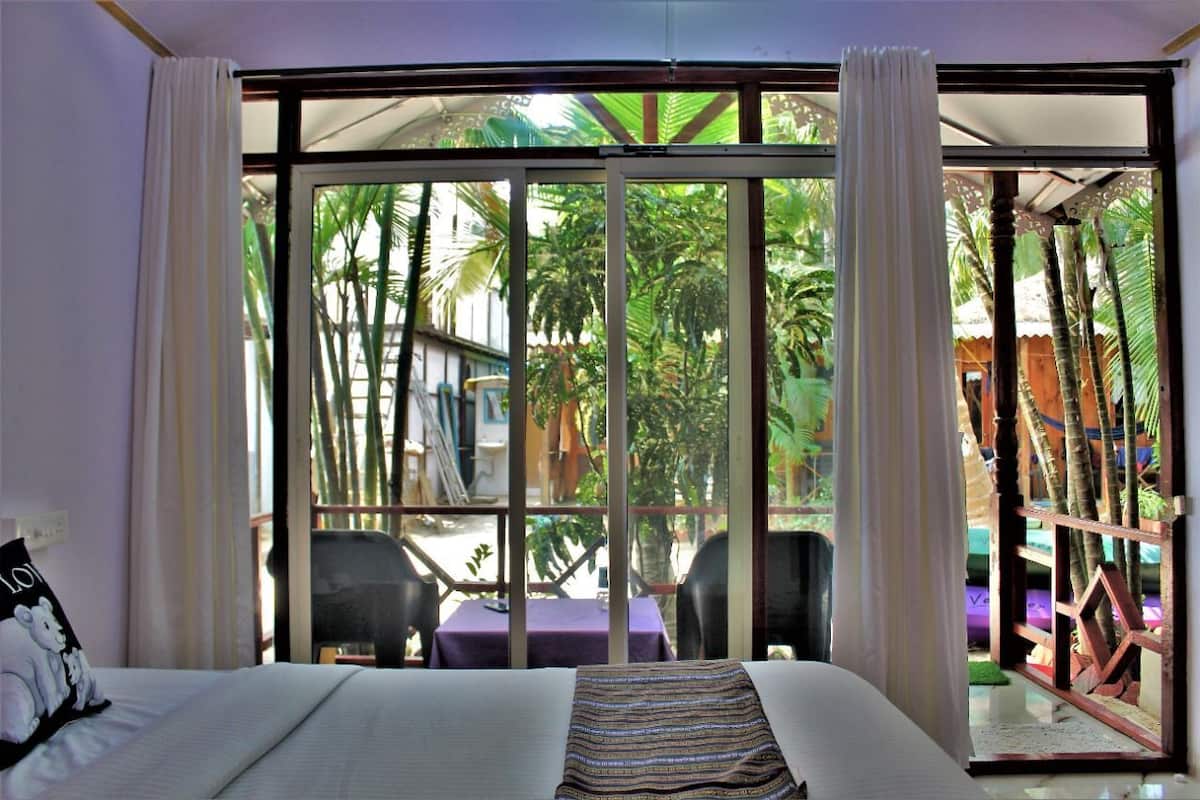 A hotel room with a large sliding glass door leading to a balcony with a view of palm trees and a building. There is a bed with a striped blanket and a pillow with a bear design.