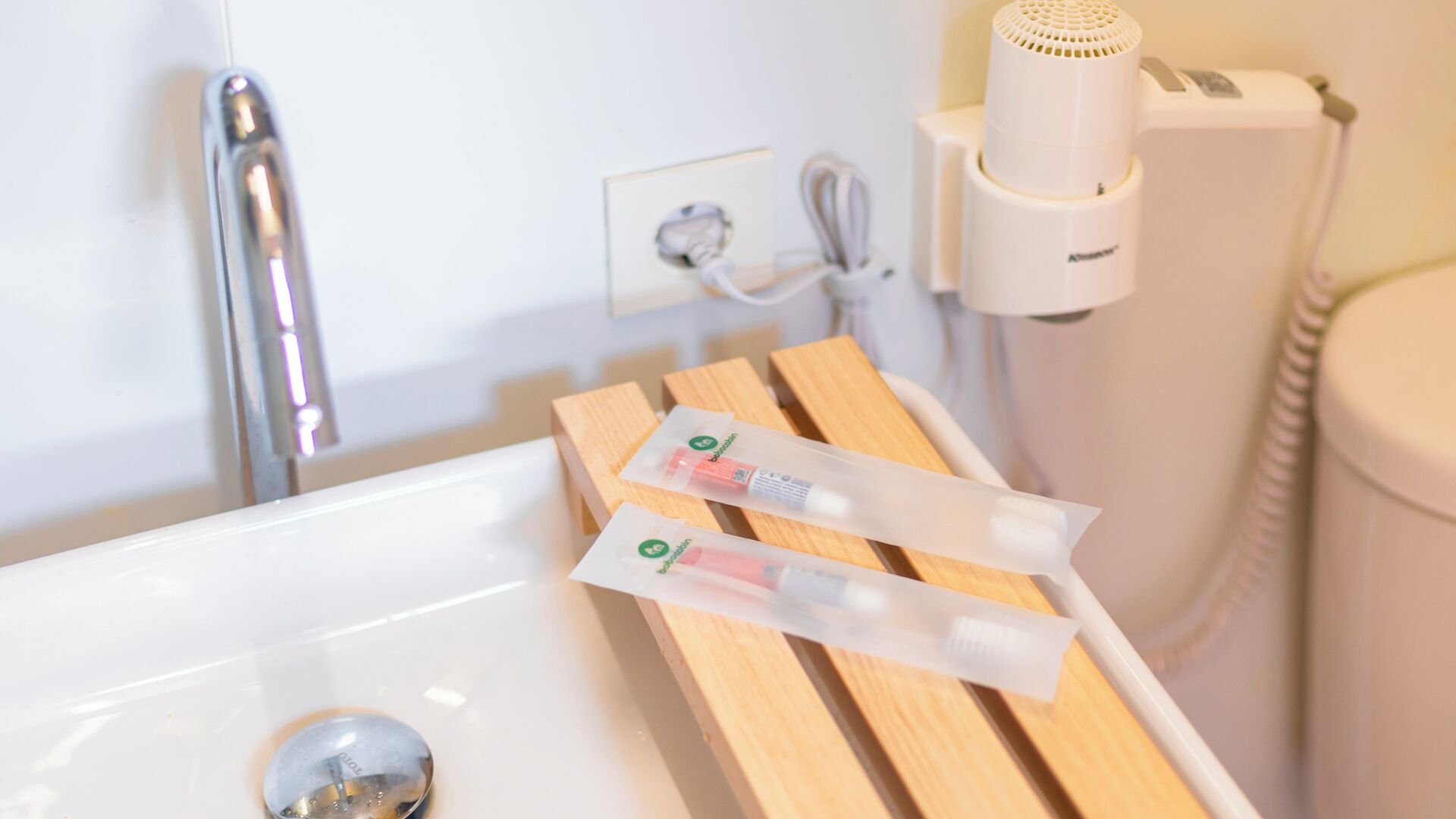 A bathroom with a white sink, wooden side table, and a hairdryer on a wall-mounted stand.