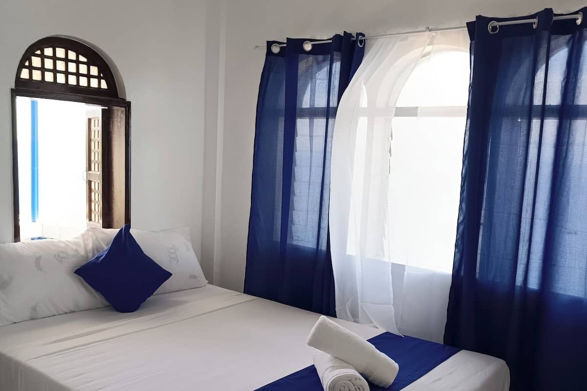 A neatly made bed with white linens and a blue accent, a window with blue curtains, and a view of the outside.