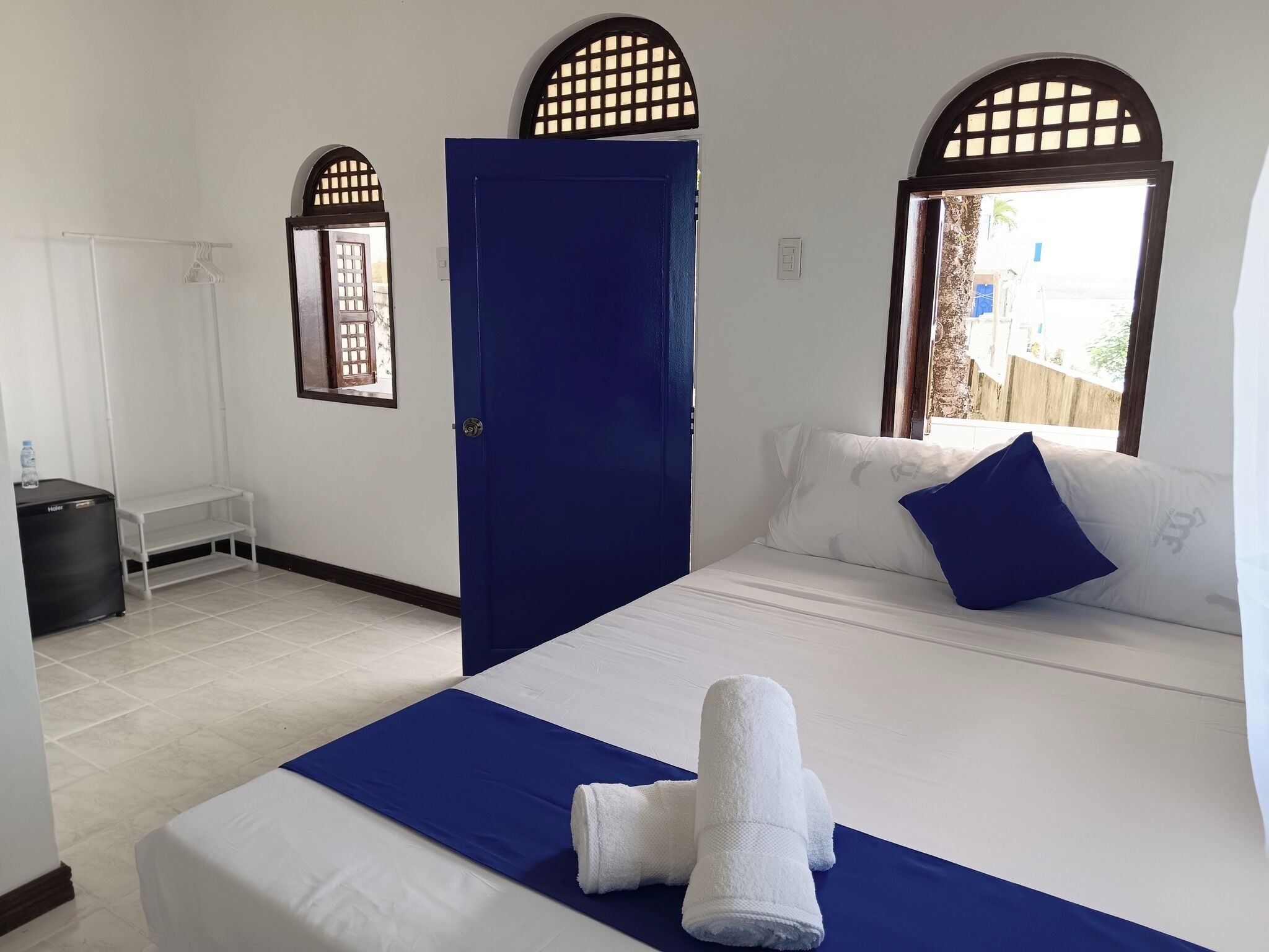 A hotel room with a bed, a blue door, a window with a view, and a small table with a water bottle.