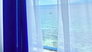 A window with sheer white curtains, offering a view of the sea and a small boat in the distance.