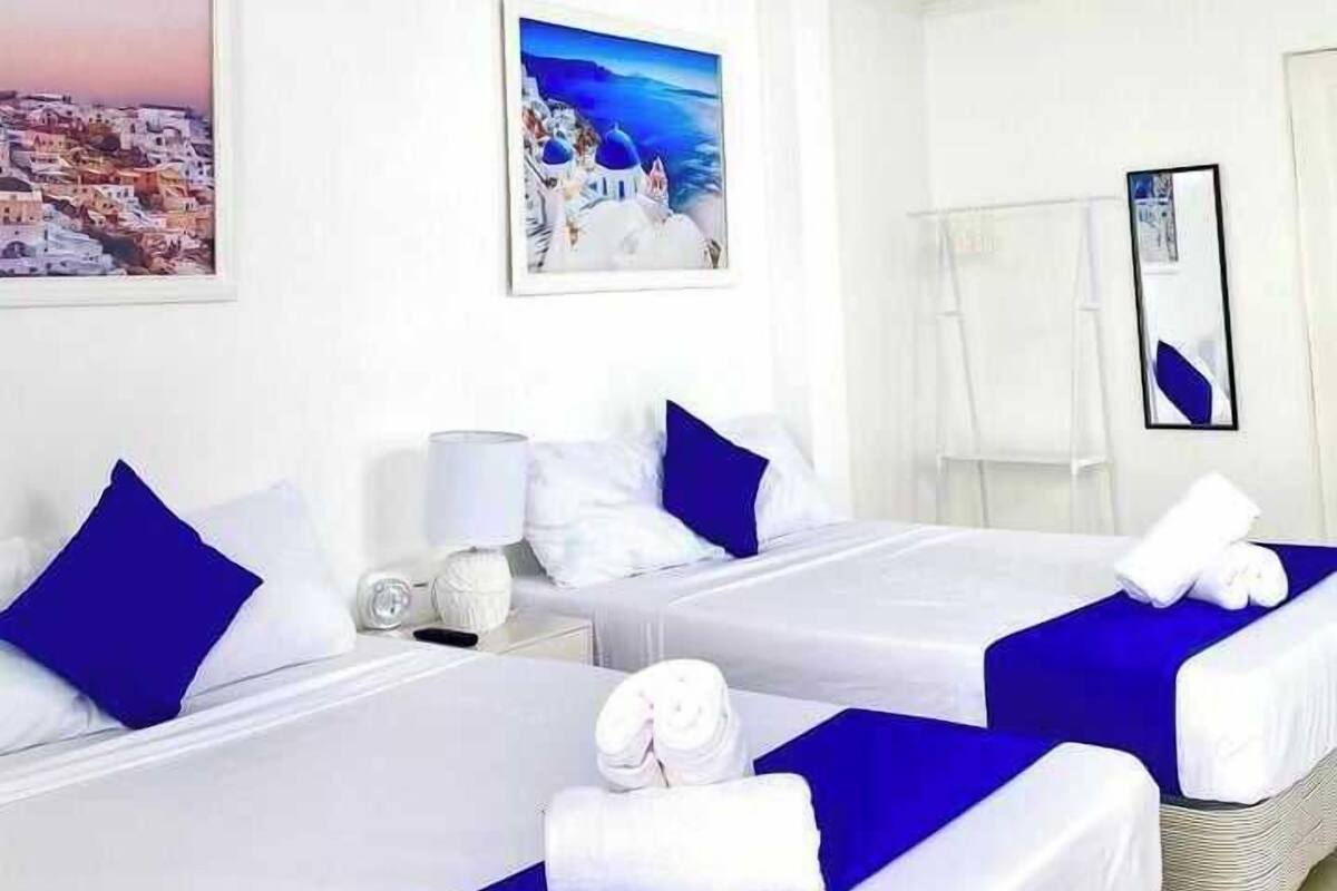 A hotel room with two beds, white bedding, blue accents, a painting of a coastal scene, a mirror, and a ceiling light.