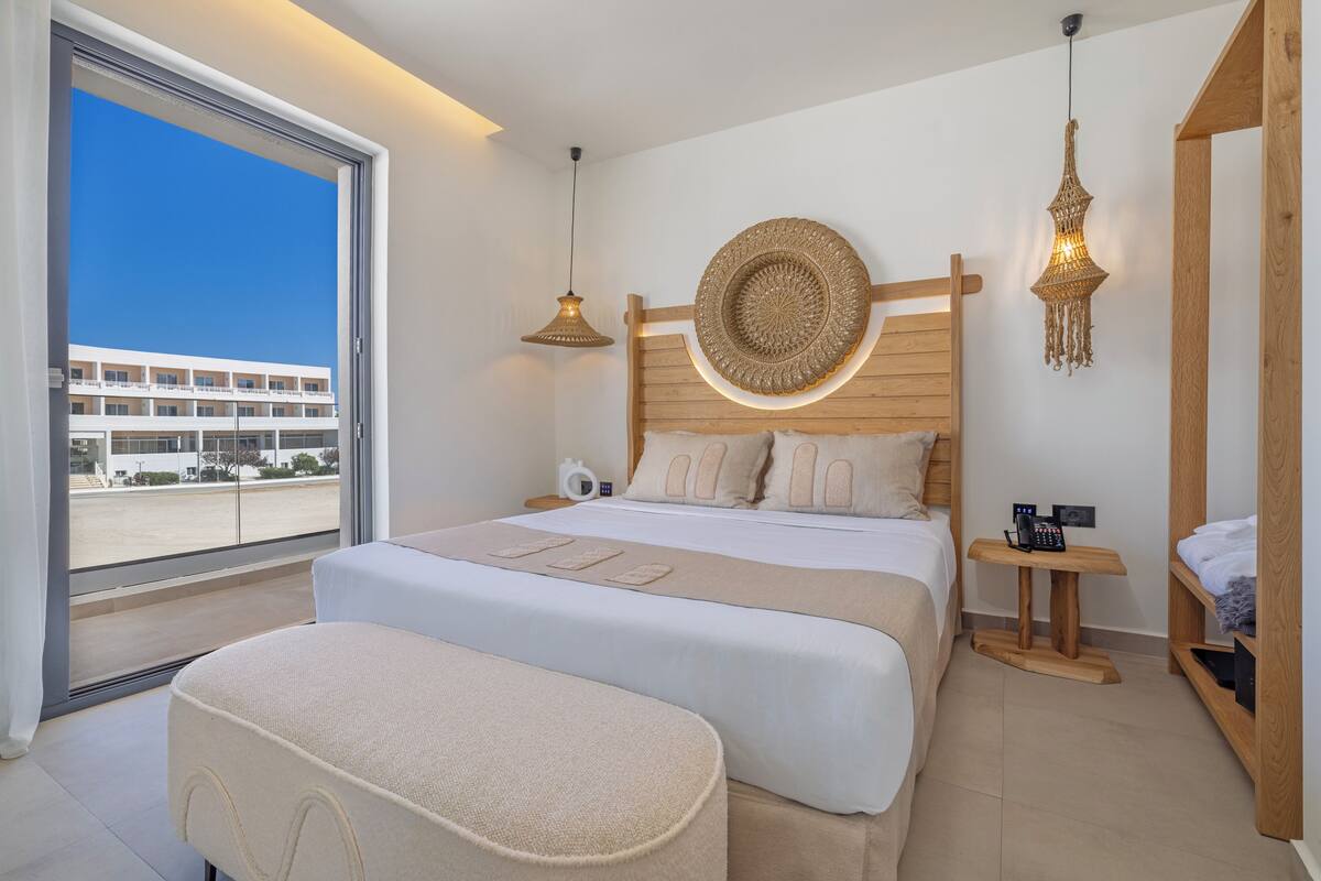 A modern hotel room with a large bed, a wooden headboard, a round decorative wall piece, a hanging light fixture, and a view of a building through a large window.