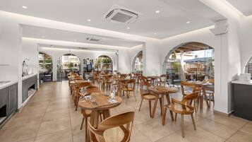 A spacious dining area with wooden tables and chairs, white walls, and large windows allowing natural light.