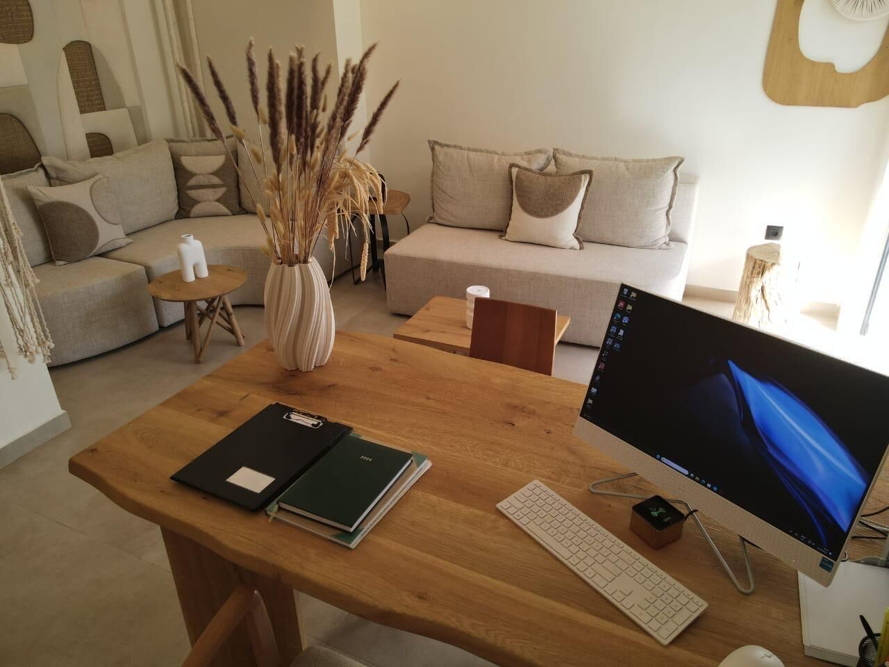 Et træskrivebord med en computerskærm, tastatur og mus, en vase med tørrede planter, samt en notesbog.