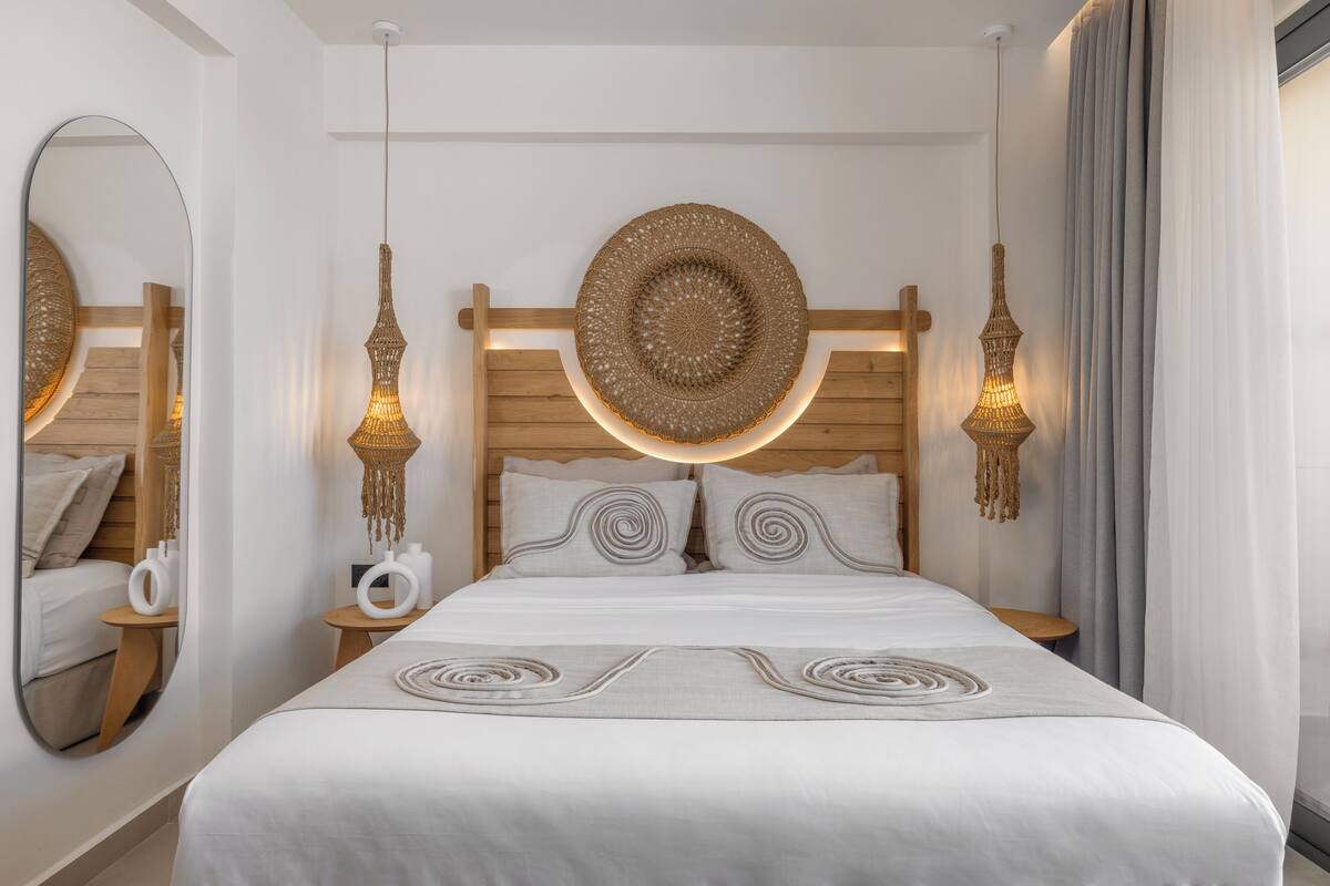 A modern hotel room with a large bed, wooden headboard, two bedside tables, a round mirror, and a large circular wall hanging.