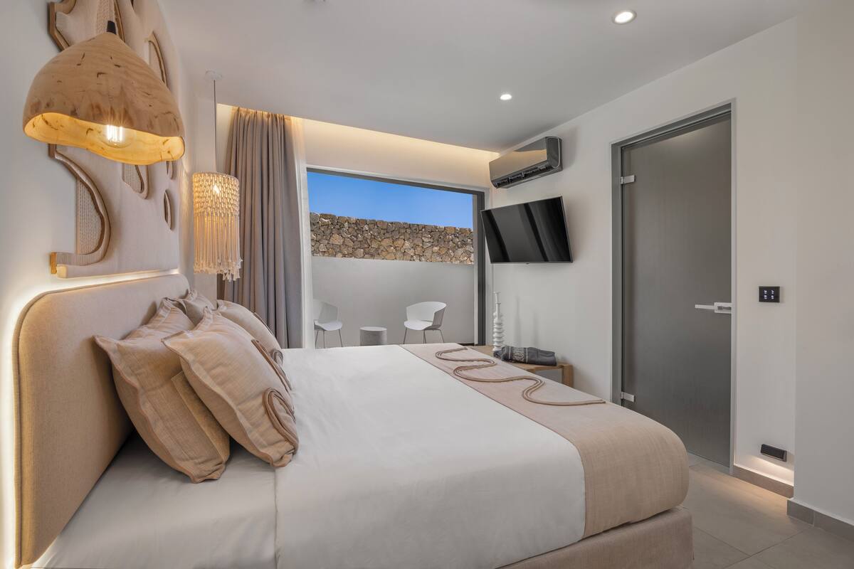 A modern bedroom with a large bed, a wall-mounted TV, and a view of a rocky landscape through a glass door.