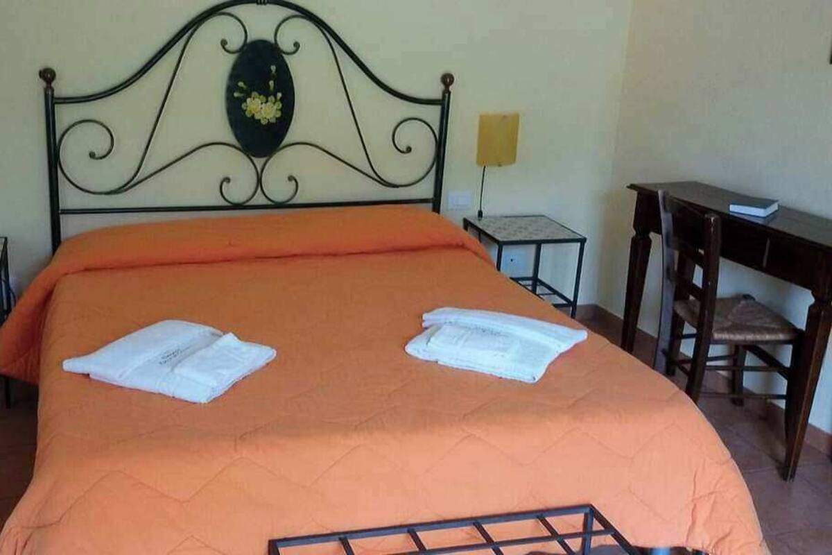 A bed with an orange bedspread, two white towels, a decorative headboard, two bedside tables with lamps, and a small desk with a chair.