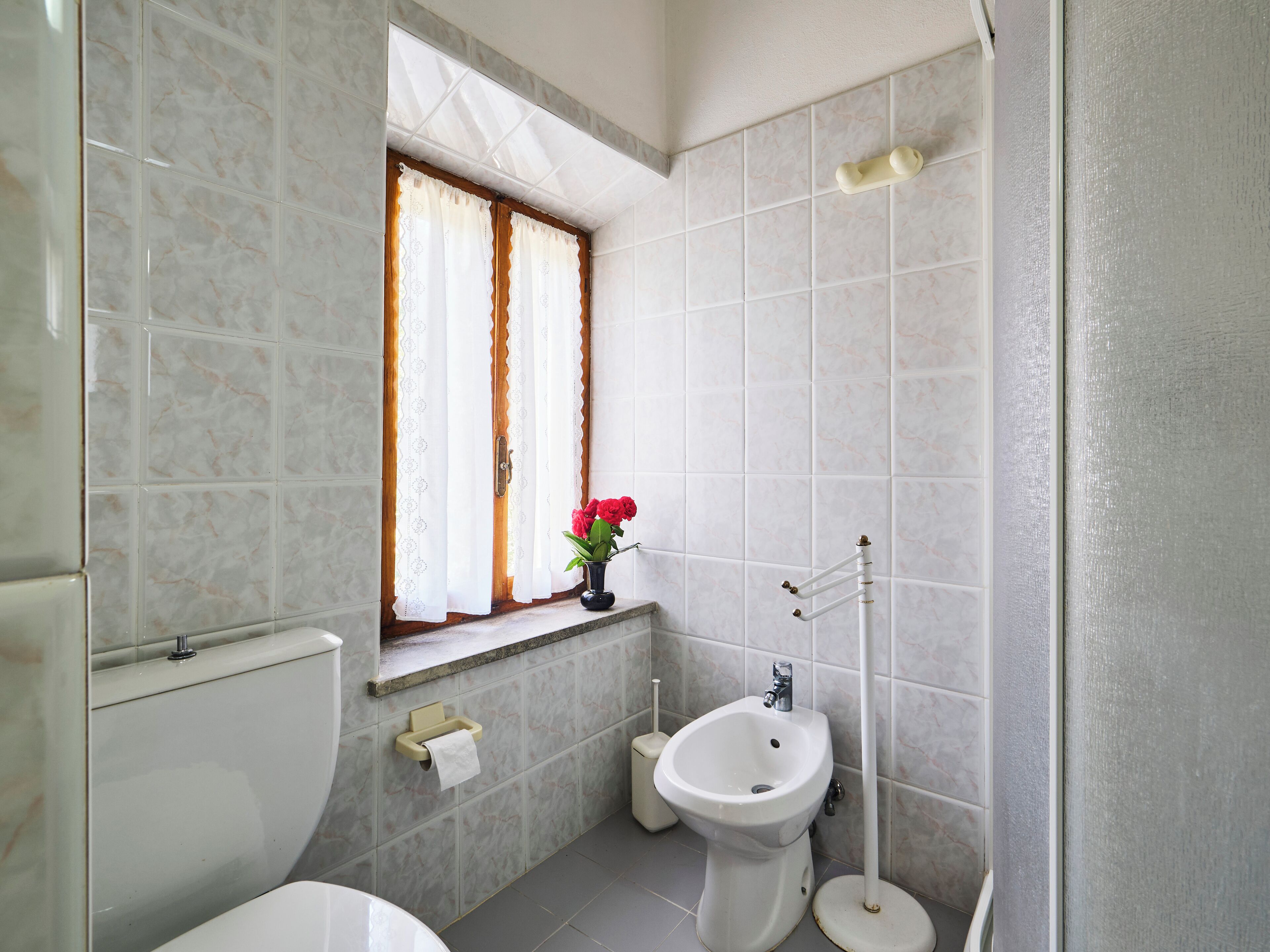 Casa de banho com sanita, bidé, lavatório, janela com cortinas e um vaso com flores.