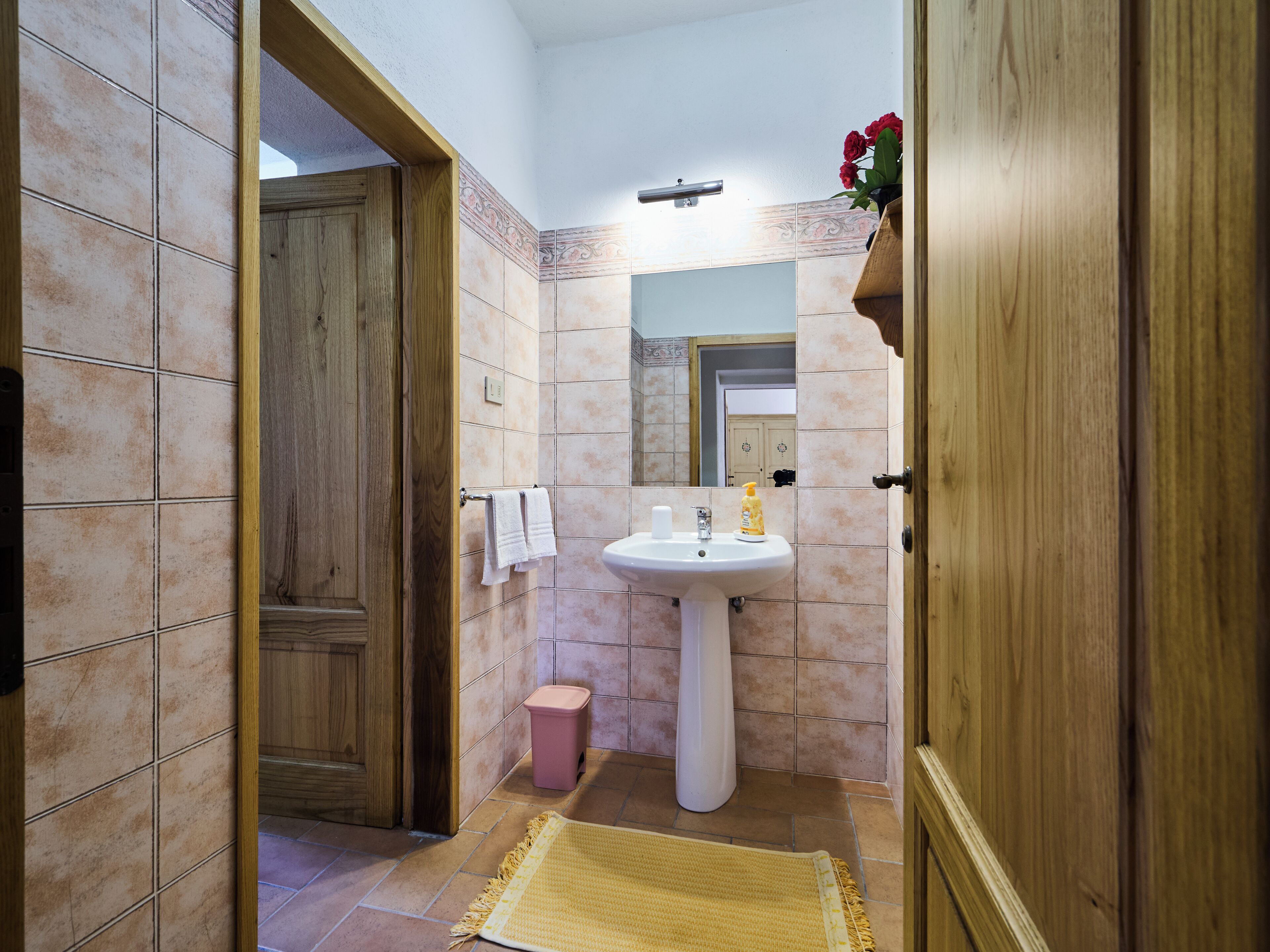 A bathroom with a wooden door, a sink, a mirror, and a small trash bin.