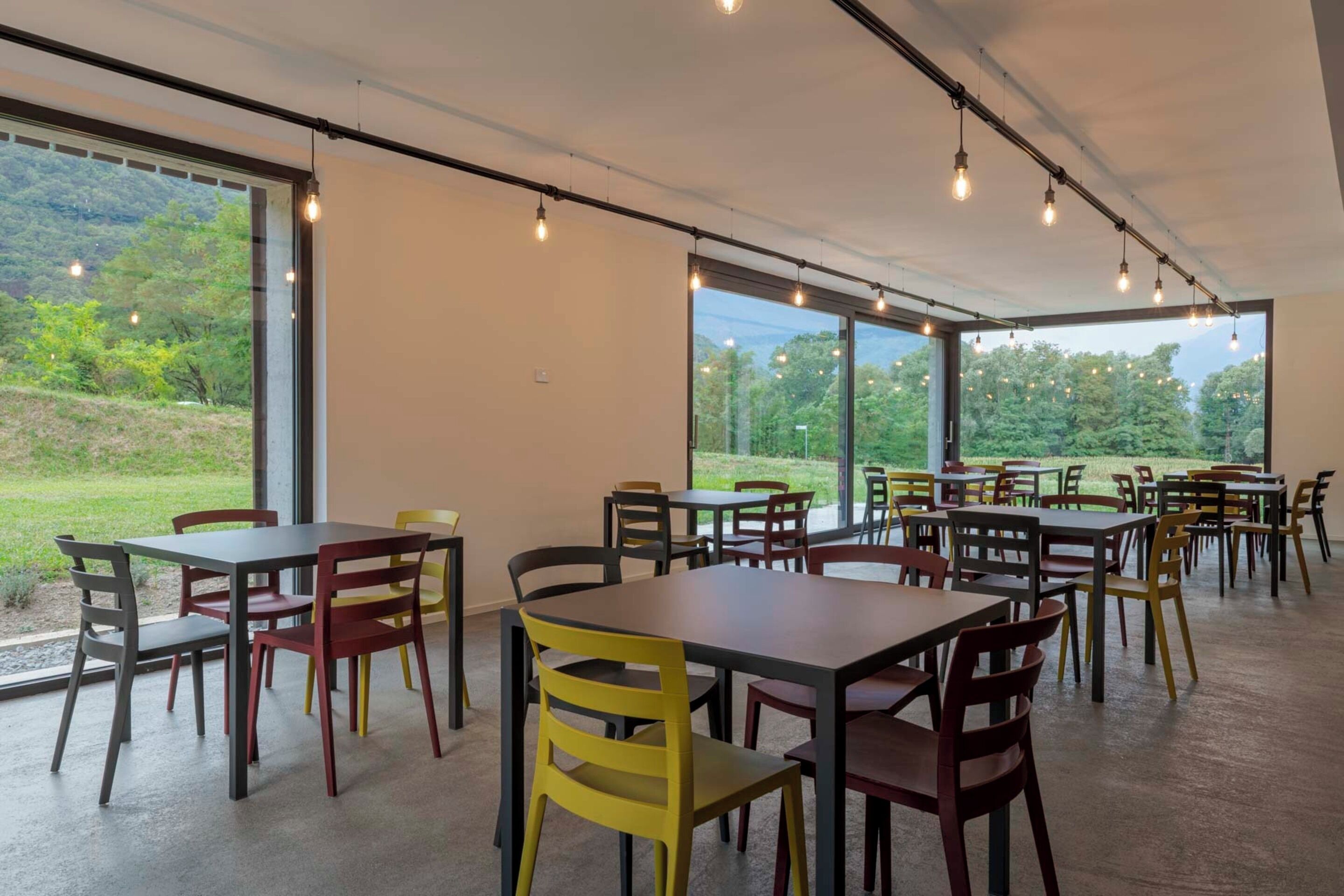 Een ruime eetruimte met meerdere tafels en stoelen, grote ramen met uitzicht op groen.
