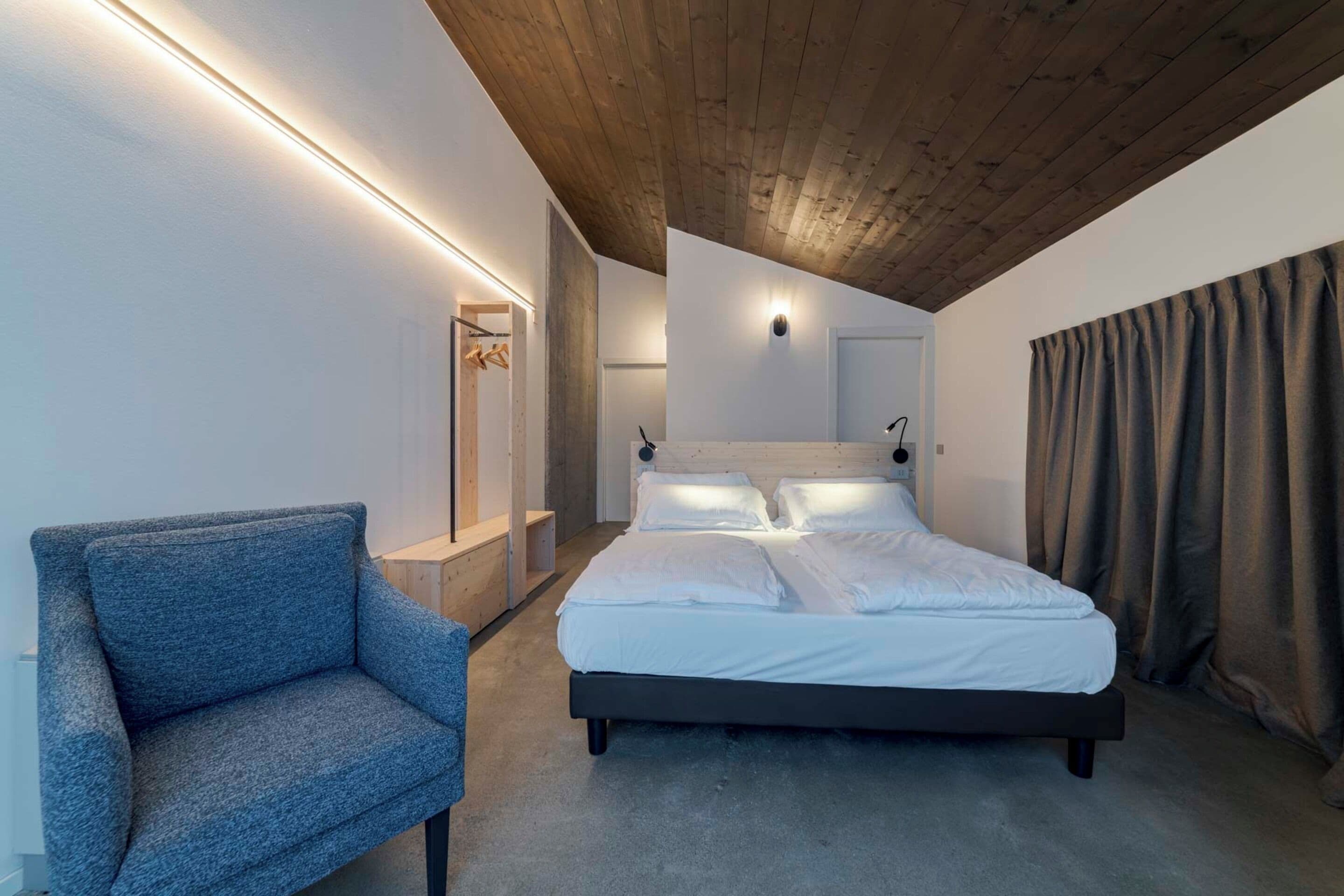 Une chambre moderne avec un plafond en bois, un lit recouvert de linge blanc, un fauteuil bleu et un banc intégré.