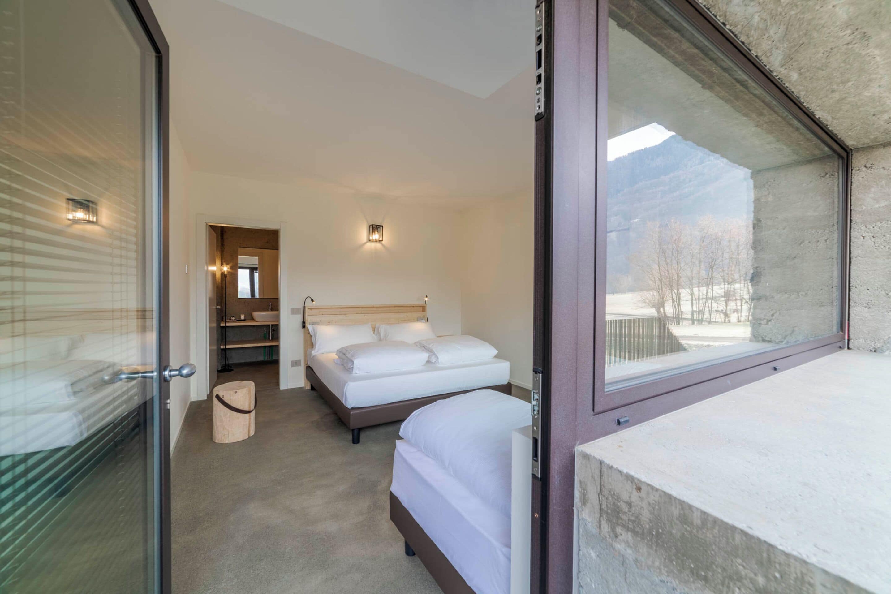 A modern hotel room with a large window, a bed, a bedside table, and a small seating area.