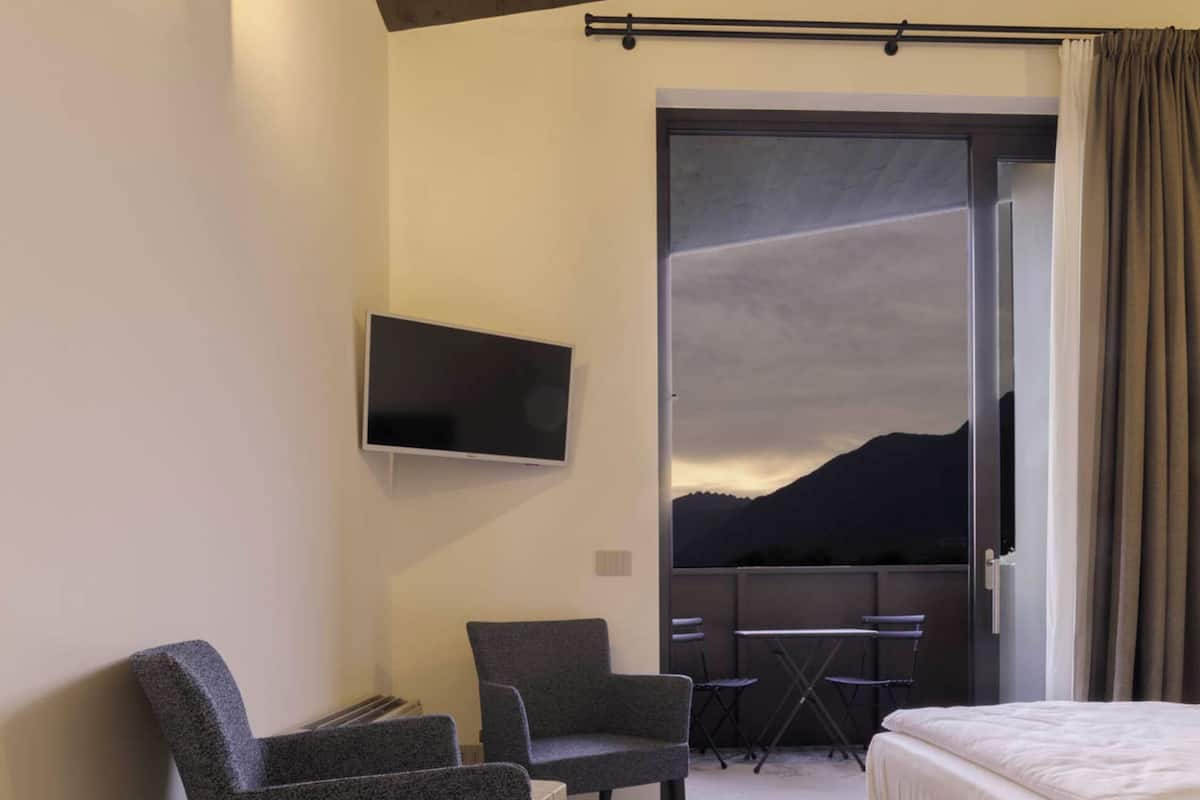 Habitación de hotel con cama, dos sillones, televisor montado en la pared y balcón con vistas a las montañas.