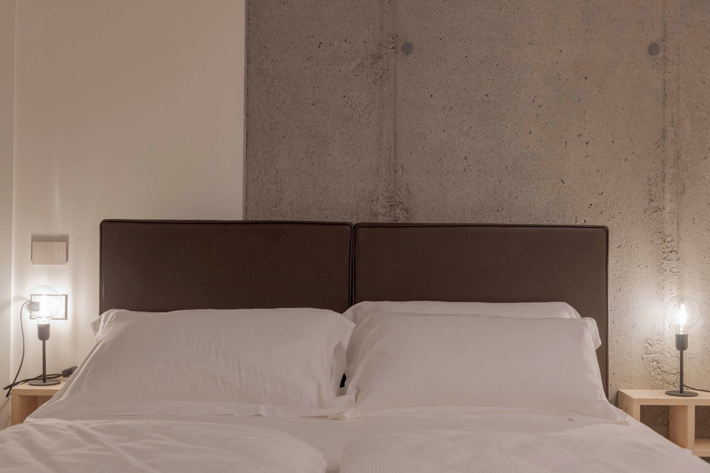 Cama com lençóis brancos, duas mesas de cabeceira com abajures e uma parede de concreto ao fundo.