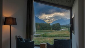Une pièce dotée d’une grande fenêtre donnant sur un paysage montagneux et une rivière. On y trouve deux chaises et un petit tabouret en bois.
