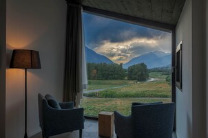 Habitación con un ventanal grande que ofrece vistas a un paisaje montañoso y un río. Hay dos sillas y un taburete de madera.