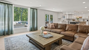 Un salon spacieux avec un canapé en cuir marron, une table basse en bois et une vue sur l’extérieur grâce à de grandes fenêtres.