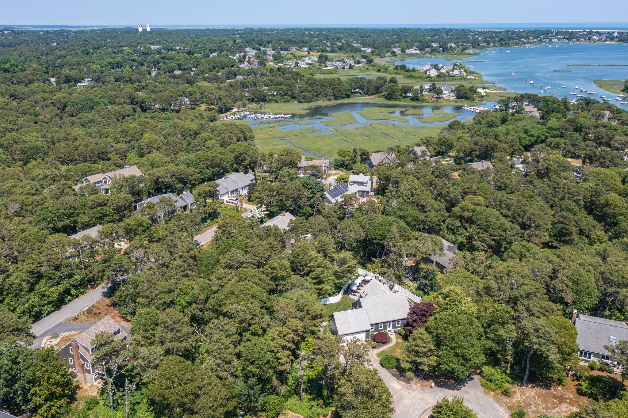 An aerial view of a residential area with houses surrounded by dense trees, a golf course, and a body of water with boats.