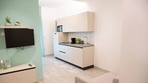 Une cuisine compacte équipée d’un four à micro-ondes, d’un évier et de placards. On y trouve une petite étagère avec des plantes et un téléviseur à écran plat fixé au mur.