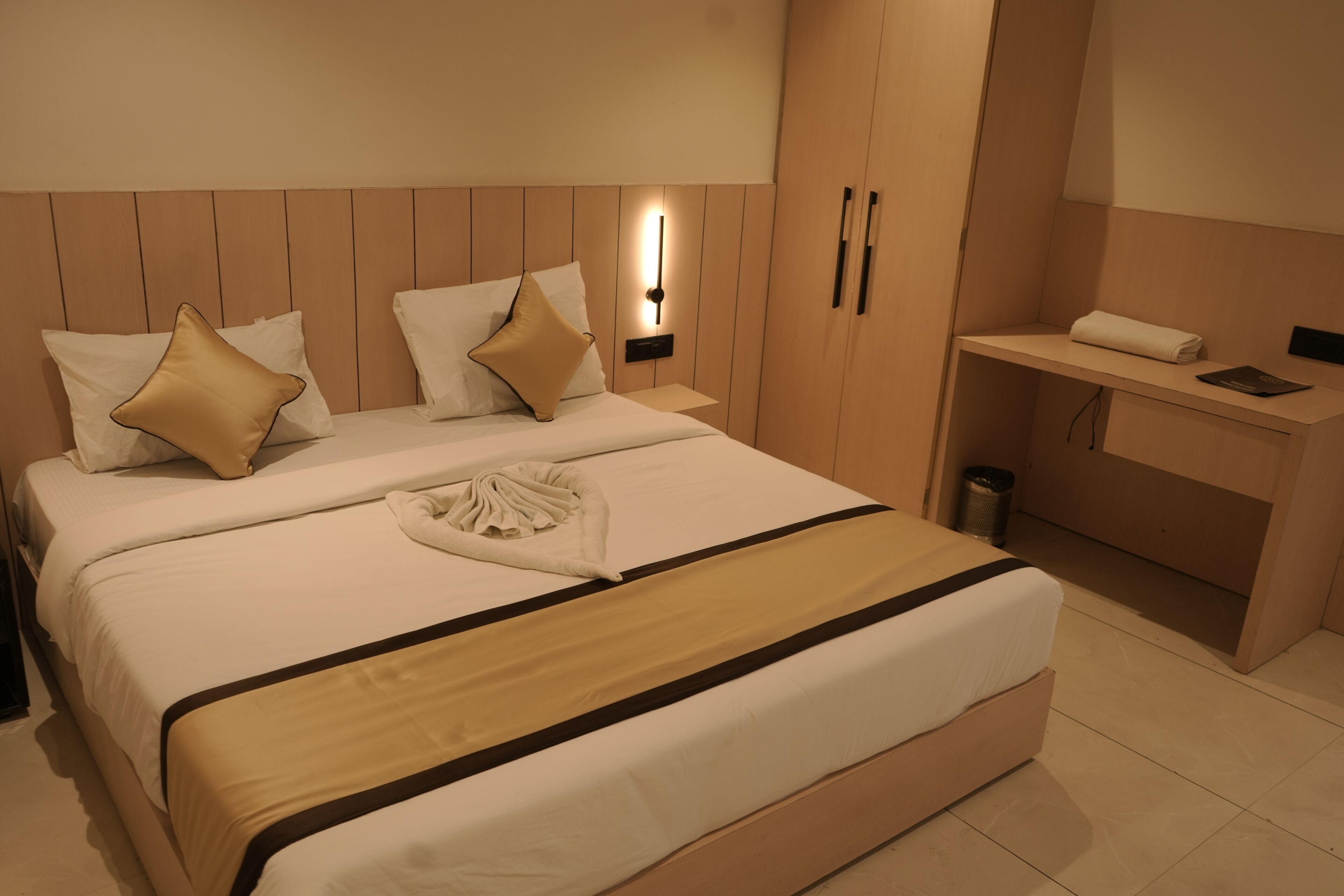 Cama bem arrumada com esquema de cores bege e branco, mesa de cabeceira com abajur e pequena área de trabalho com toalha e um livro.