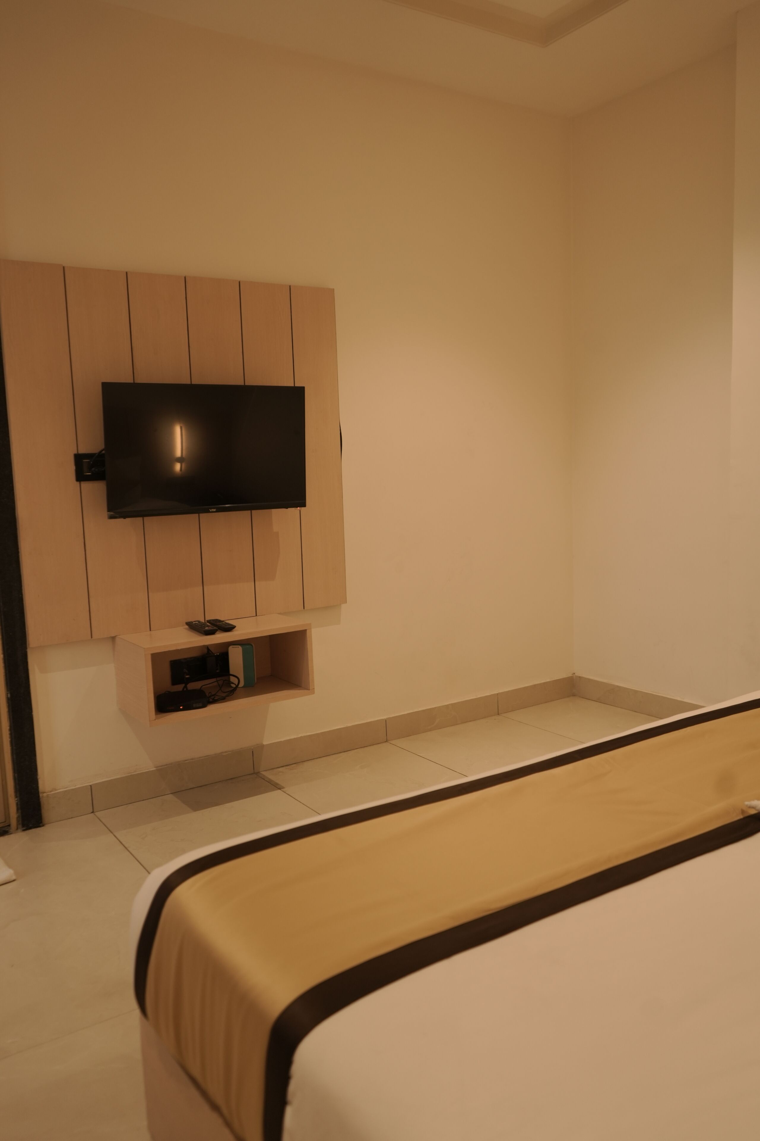 A hotel room with a bed, a TV mounted on the wall, and a small shelf with electronic devices.