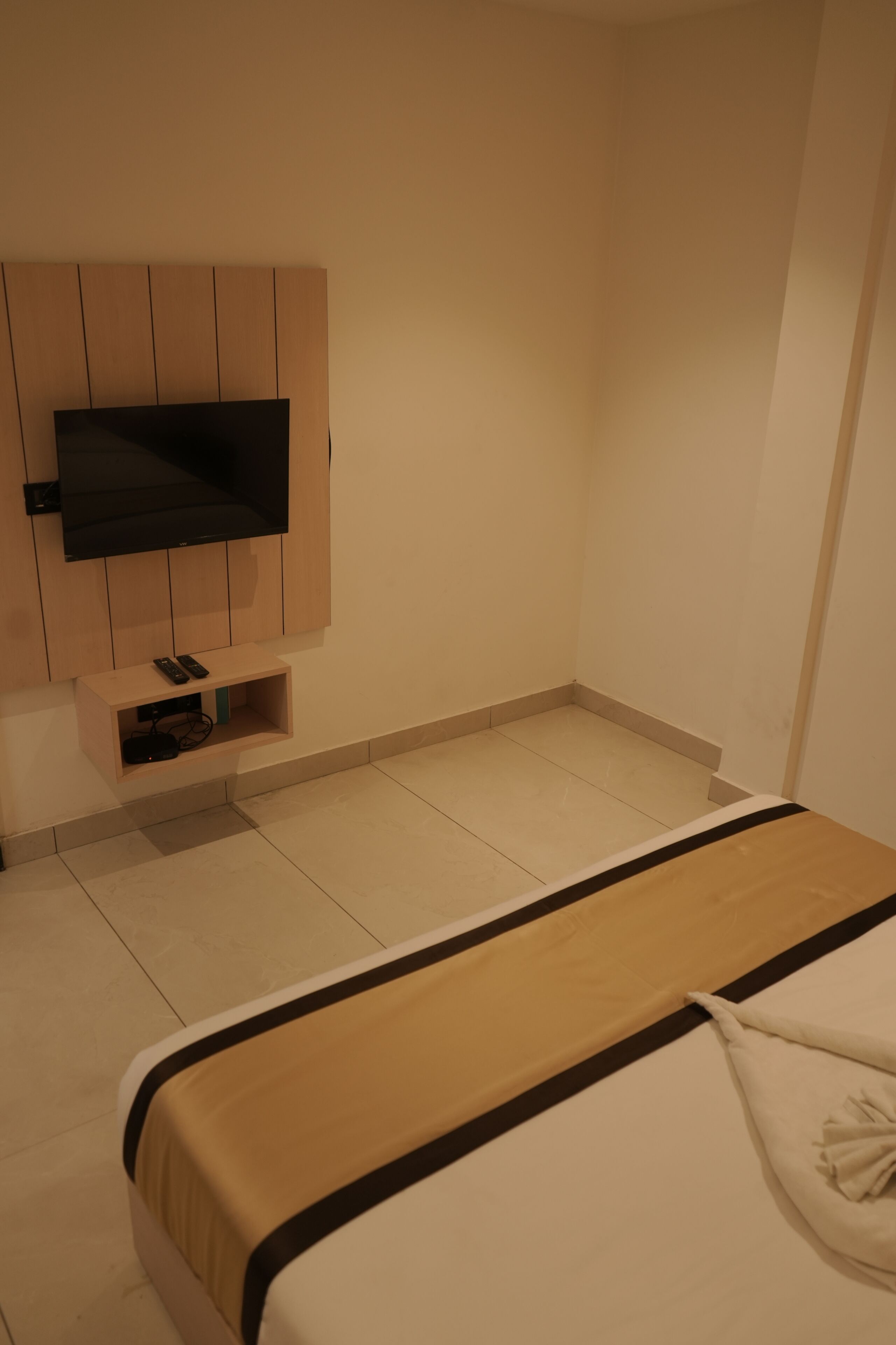Une chambre d’hôtel avec un lit, un téléviseur fixé au mur, une table de chevet et une petite étagère avec une télécommande.