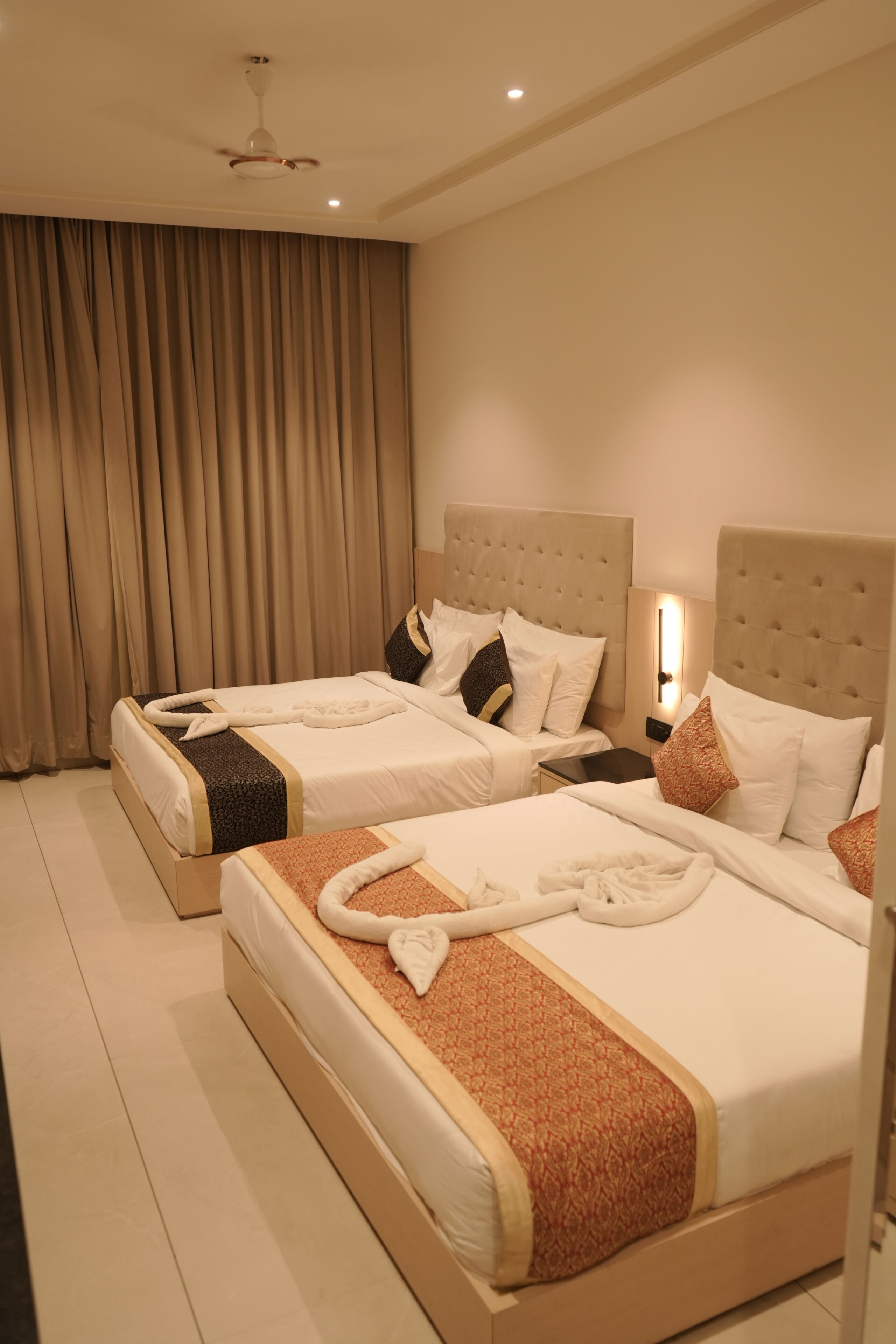 A hotel room with two beds, beige curtains, a ceiling fan, and a nightstand with a lamp.
