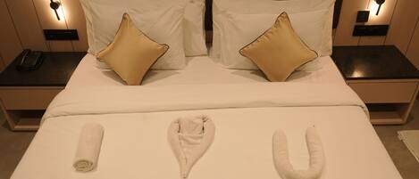 Una cama bien hecha con dos almohadas blancas y dos almohadas color beige, una toalla enrollada y una toalla doblada en forma de 'U' en una cama de hotel.