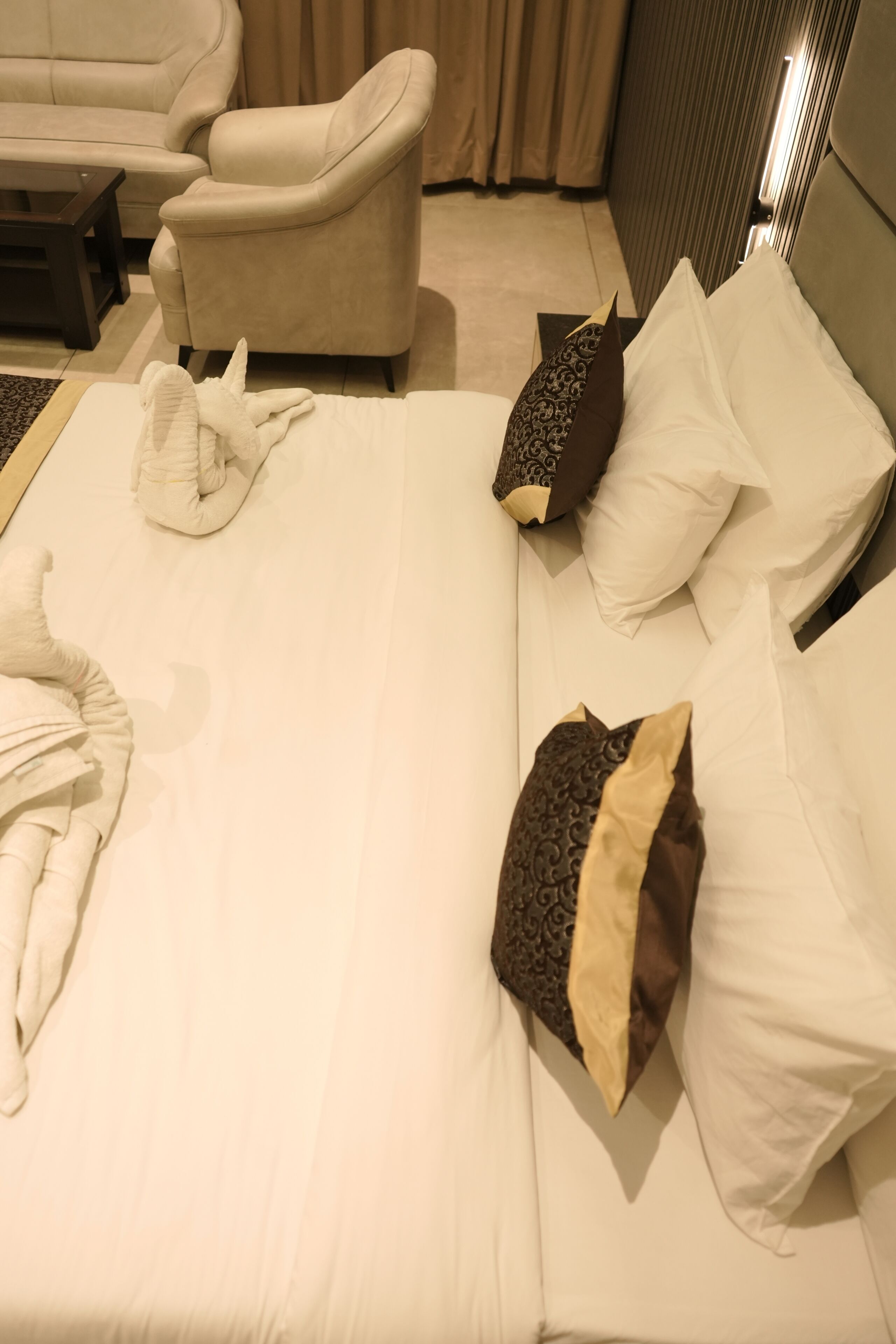 A neatly made bed with white and patterned pillows, a beige armchair, and a small table in a hotel room.