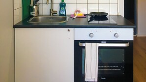A compact kitchen with a sink, stove, oven, and a shelf above holding pots and a bowl.