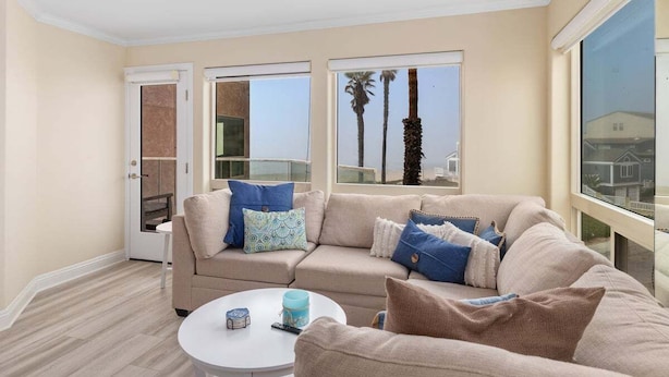 Ein Wohnzimmer mit einem beigen Sofa, einem runden Couchtisch und zwei Fenstern mit Blick auf Palmen und ein Haus.