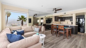 Ein modernes Wohnzimmer mit einem beigefarbenen Sofa, einem Couchtisch und einem Essbereich mit Holzsesseln und einem kleinen Tisch. Im Hintergrund ist eine Küche mit dunklen Schränken und Edelstahlgeräten zu sehen.