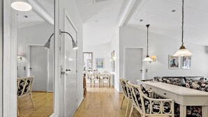Un pasillo con paredes blancas, suelo de madera y una zona de comedor con mesa y sillas blancas. Hay lámparas colgantes sobre la mesa del comedor y un sofá con tapicería floral en la zona de estar.