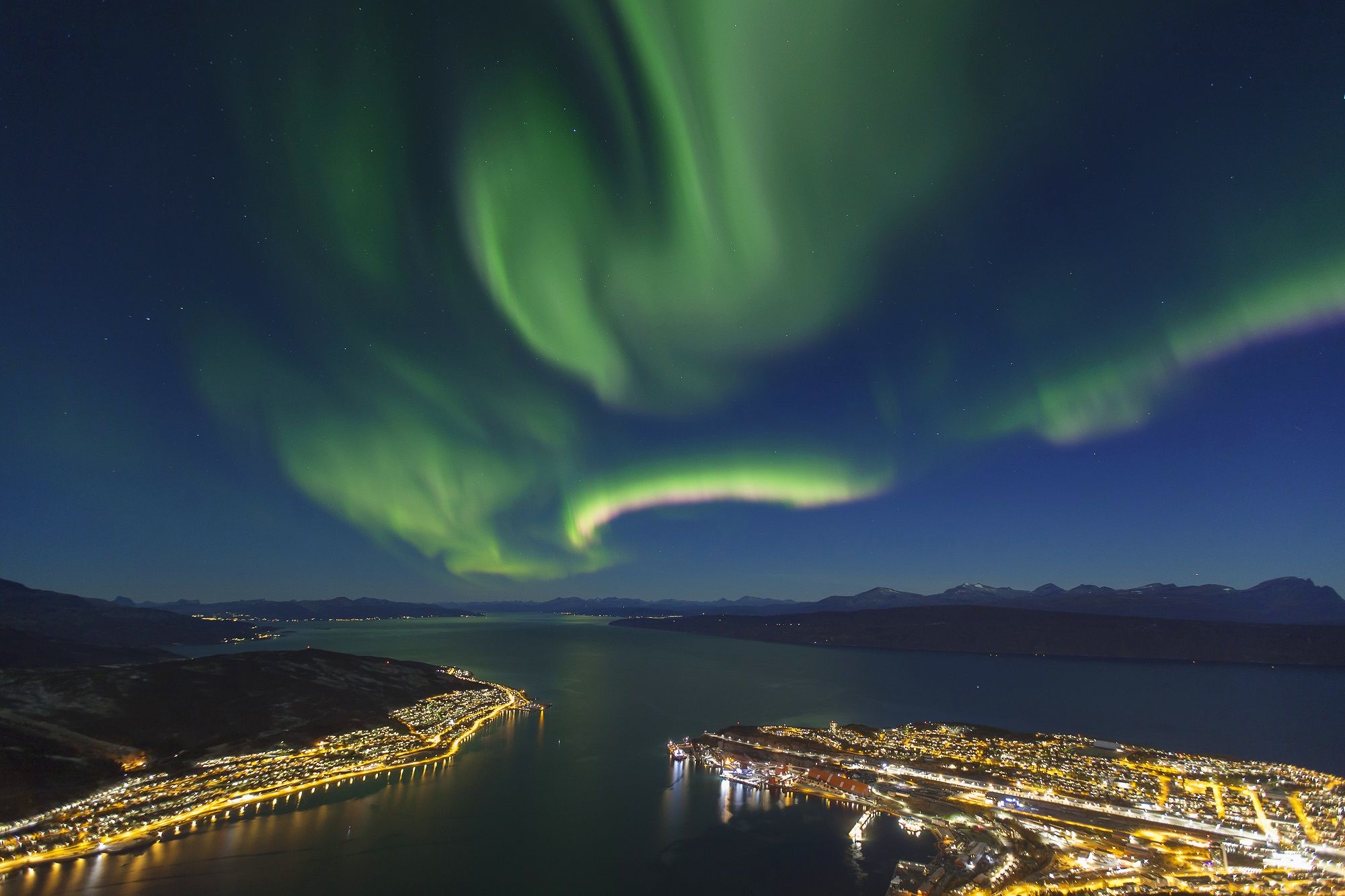 Vista nocturna de una ciudad costera con un río, edificios iluminados y auroras boreales.